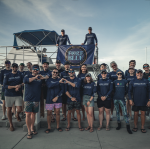 FORCE BLUE team members gathered in front of a dive boat, wearing FORCE BLUE shirts and posing beneath the organization’s flag at a marina.