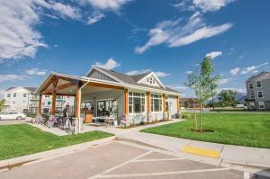 The Village on 4th leasing office and clubhouse exterior in Springville, Utah, showing residents gathered on a covered patio next to parked bicycles and a green lawn.