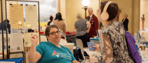 Person wearing an Autism Foundation of Oklahoma shirt talks to a visitor with headphones at an indoor event table with certificates displayed behind.