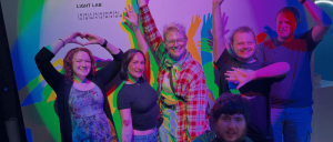 Group of six people posing cheerfully in colorful lighting at the Light Lab with shadows cast on the wall behind them.