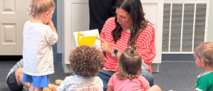Woman reading a book with a yellow duck illustration to a group of young children seated on the floor in a classroom.