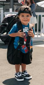 Young child wearing a backpack and holding the straps while standing outdoors at a community event.