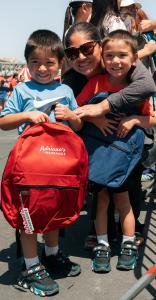 A mother smiles with her two children as they proudly hold free Adriana’s Insurance backpacks during a community Back-to-School event.