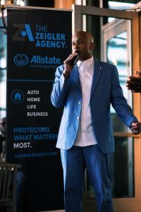 Chris Zeigler of The Zeigler Agency speaking into a microphone beside an Allstate agency banner at a professional event.