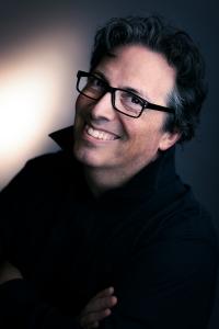 Headshot of a smiling man with black-framed glasses, dark wavy hair, and a dark collared shirt looking toward the camera.