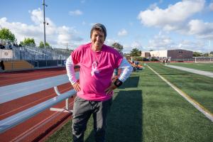 Stuht stands with her hands on her hips beside the bleachers on a school athletic field, wearing a bright pink T-shirt over a white long-sleeve shirt.