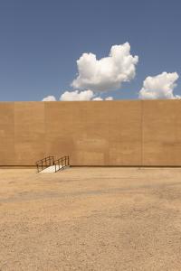 A contemporary photograph of a bridge transitioning from a dirt lot straight into a tall brown wall, with clouds above in a blue sky.