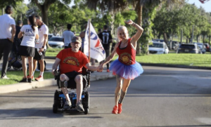 Devil Dog 5K/10K participants hold hands on the racecourse, including a veteran using a motorized wheelchair and a runner wearing a patriotic tutu.