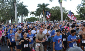 A large crowd of Devil Dog 5K/10K participants gathers beneath palm trees while runners hold American flags.