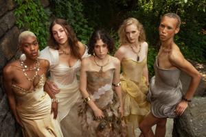 Five models wear earth-toned designs from Mikael Pono’s Genesis collection, featuring natural fibers, organic materials and nature-inspired textures, posed outdoors amid rocks and lush greenery.
