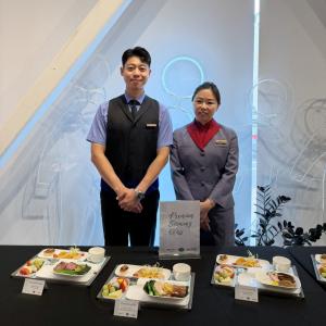Two China Airlines cabin crew members stand behind a display of Premium Economy Class in-flight meal selections, including a featured Wenwen collaboration dish.
