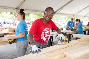 A smiling volunteer in a red Bunks Across America T-shirt sands a wooden board under a work tent, with other volunteers sanding in the background.