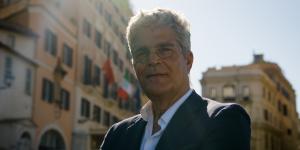 Elio Epifani smiling outdoors on a Rome street, wearing glasses and a dark blazer, with historic buildings behind him