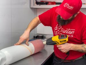 Two J. Blanton Plumbing technicians in red uniforms pour epoxy resin into a felt pipe liner inside a service trailer, preparing for trenchless sewer pipe repair.