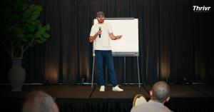 Nikolaj Kloch holding a microphone on stage beside a whiteboard, speaking to an audience at a business conference.