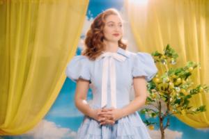 Singer-songwriter Ashley Ann in a pale blue dress with a white bow, standing between yellow curtains beside a lemon tree.