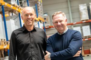 Hayco van Gaal and Noah Rogers stand side by side in a warehouse at RPI Print's Eindhoven plant, both smiling at the camera. Van Gaal, on the left, wears a black button-up shirt. Rogers, on the right, wears a navy sweater over a checked collared shirt, wi