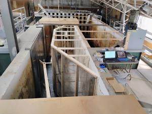 Indoor hydraulics lab with a large concrete water channel divided by wooden frames and mesh screens, next to a workbench with two laptops, one showing an underwater camera view.