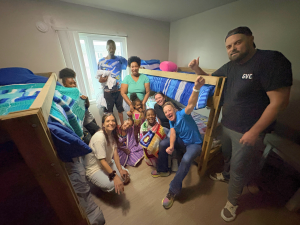 A group of SHP volunteers and family members smiling and posing around two newly delivered bunk beds in a bedroom.
