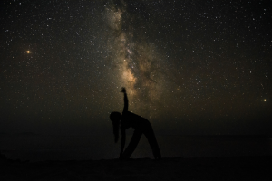 Silhouette of a person practicing yoga beneath a star-filled night sky, with the Milky Way visible overhead, illustrating star bathing, mindfulness, and dark-sky wellness tourism.