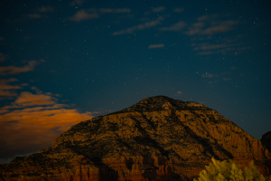 Sedona, Arizona, red rock mountain illuminated by warm evening light beneath a deep blue, star-filled night sky with scattered clouds, highlighting the region's scenic dark-sky landscape.