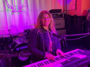 Image of Sofia Nunzia, a teenage girl with curly blonde hair sitting at a keyboard and microphone with a Yamaha drum set behind her