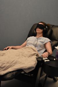 A woman with short wavy hair wearing a black eye mask, headphones and a pale blue short sleeves blouse sits in a black recliner under a grey blanket for ketamine infusion therapy. She looks very relaxed,