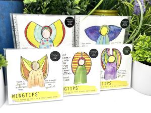 Five WingTips packages are displayed on a white shelf among houseplants. The top art print within each set is visible through clear, plastic packaging, with a bright and colorful angel image central to each piece, surrounded by handwritten angelic advice.