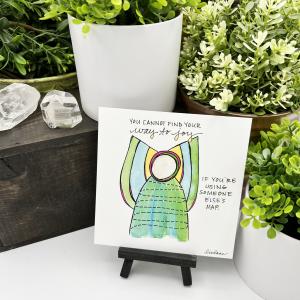 A WingTips print is displayed on a on a white shelf, surrounded by green houseplants and large clear crystals. The whimsical angel image at the center of the print is wearing a bright green dress with what appears to be a black and white ribbon winding in