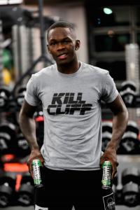 Fresh off the most successful launch in company history with Joe Rogan’s Flaming Joe, Kill Cliff, the undisputed clean energy Champion of America and Beyond, is launching the best Fight Club in the world, headlined by current UFC Middleweight Champion Israel Adesanya Israel Adesanya in a Kill Cliff shirt with Kill cliff beverage cans in a gym