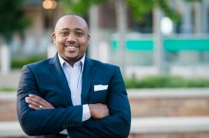 Dontaye Carter in dark blue suit with no tie and a light blue shirt with arms folded while smiling.