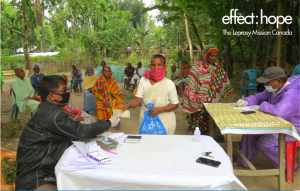 Photo: In a cleared area surrounded by trees, two administrative tables are set up. Project participants in Bangladesh line up to receive assistance during COVID-19 pandemic.