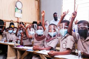 Young students wearing masks and face shields sitting at their desks and raising their hands.