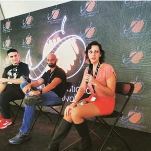 Koura Linda, Trent Duncan, and another filmmaker sit on black folding chairs in front of an LED wall with festival logos on it.  Koura is wearing an orange mini skirt, pink top, and black knee-high boots, with dark hair, holding a microphone.