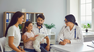 A female pediatrician talking to a family of a mother, father and young girl in a medical office. The doctor is taking notes and the young girl is holding a stuffed animal.