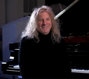 Composer David Arkenstone at his piano in a black sweater, with long white hair