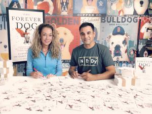 Dr. Becker and Rodney Habib seated at a desk signing bookplates for The Forever Dog