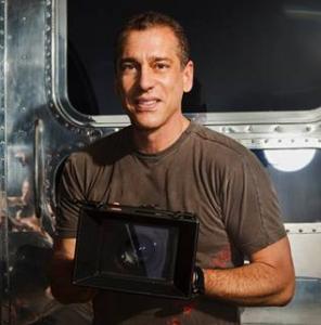 Michael Grecco faces the camera and smiles, while holding a large, square, black camera