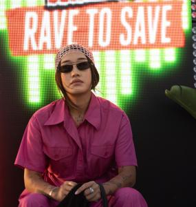 Peggy Gou sitting in front of a Rave to Save sign in a pink jumpsuit, headband and sunglasses
